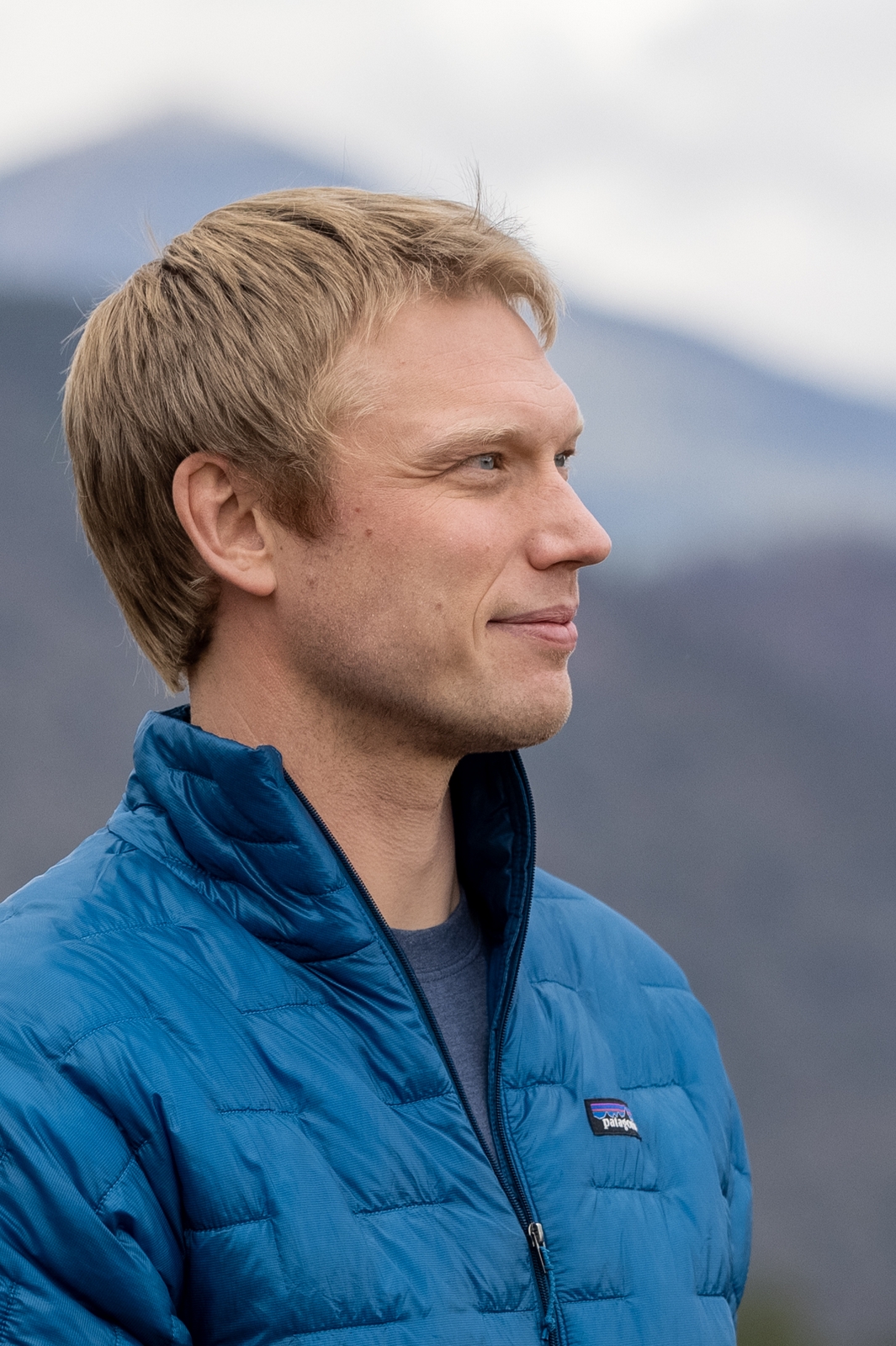 A man with blond hair looks into the distance. He is wearing a blue down coat.