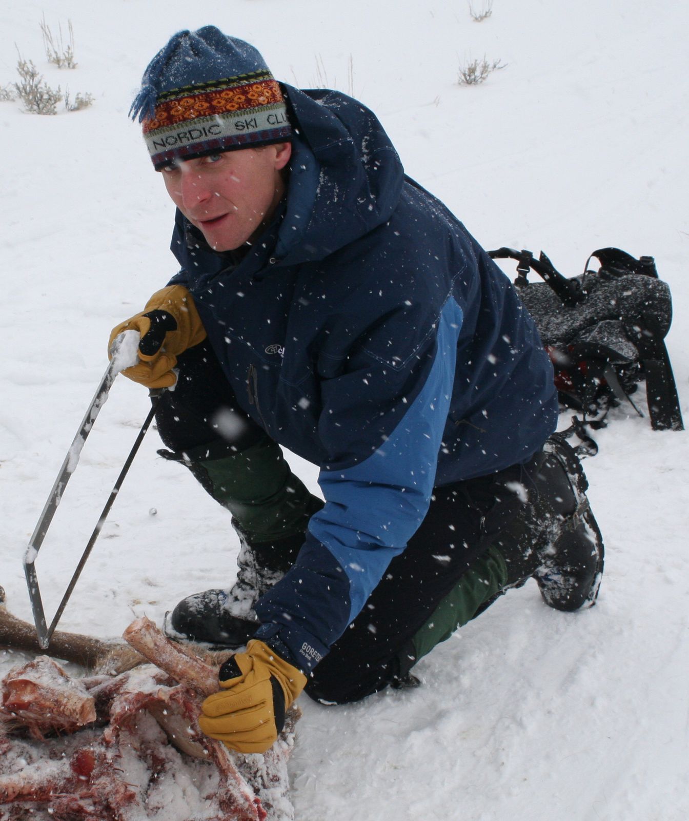A man kneels in the snow holding a saw and an animal carcass.