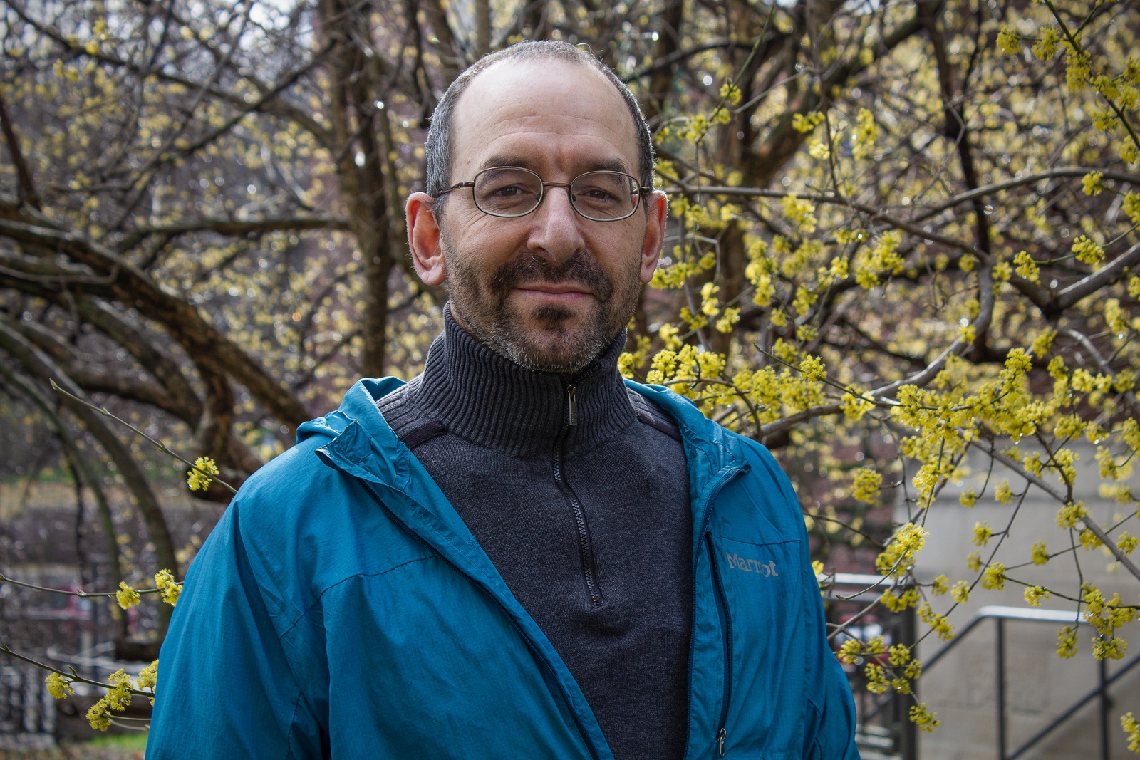 A man with dark hair and glasses stands in front of a tree with yellow blossoms. He is wearing a blue turtleneck sweater and blue raincoat.