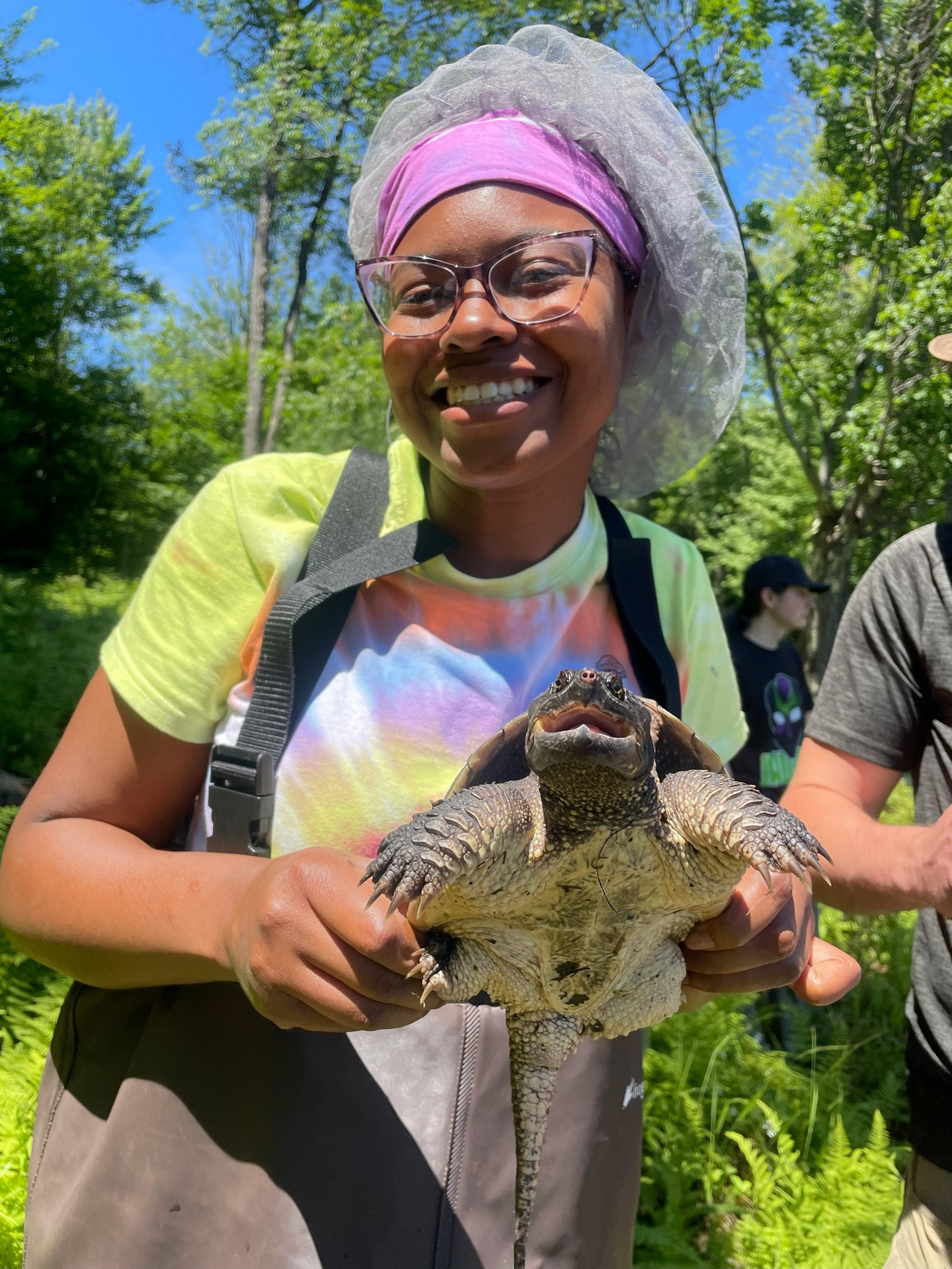 A woman with brown hair in a ponytail and sunglasses on her head, wearing a purple jacket. Mountains are in the background.
